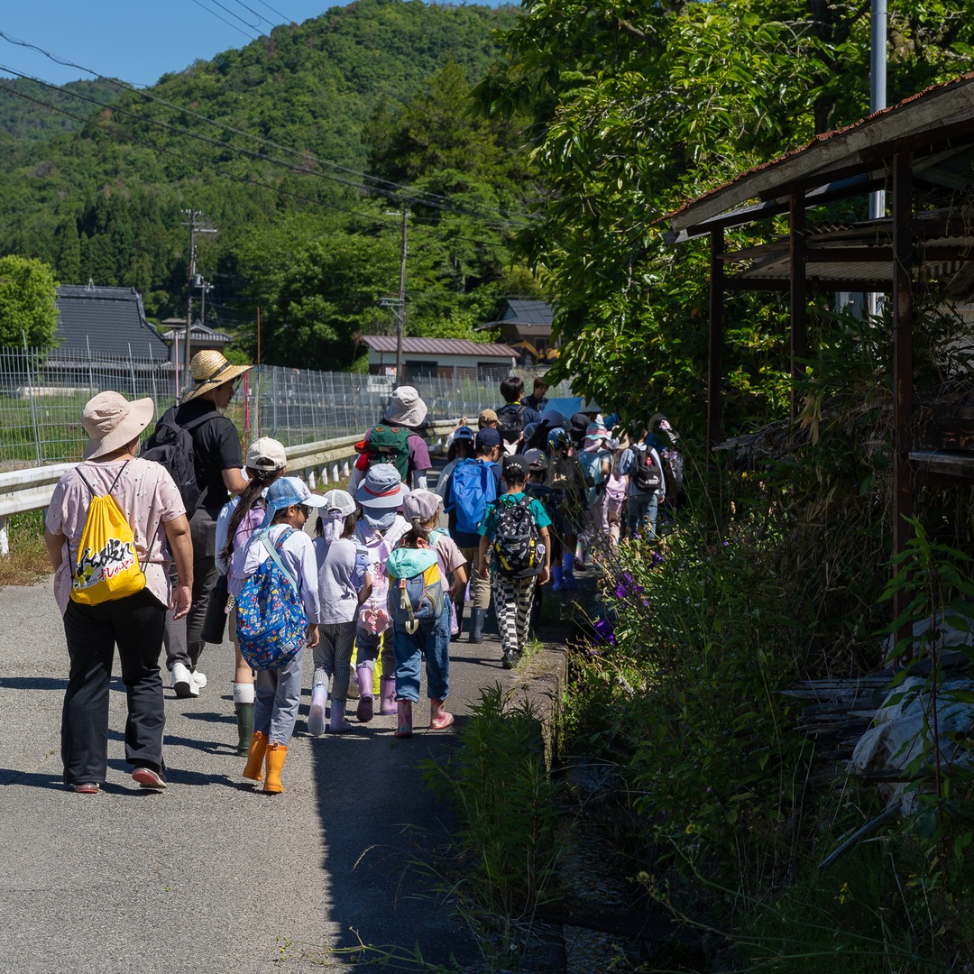 里山の道を一列で歩いて古民家へ向かう参加者たち